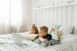 Dzieci leżą na łóżku, oglądając edukacyjne bajki dla dzieci na laptopie.