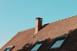 Brązowy komin na dachu z dachówki, pod błękitnym niebem. Może być mokry od deszczu, ale wygląda na suchy.