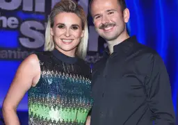 Uśmiechnięci Sławomir i Kajra pozują do zdjęcia na tle niebieskiej kurtyny. Kajra ma na sobie błyszczącą sukienkę, a Sławomir czarną koszulę.
