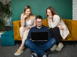 Trzy osoby pracują przy laptopie. Dwie kobiety siedzą na kanapie, a mężczyzna na podłodze. Zastanawiają się, cro co to jest.