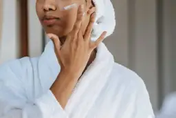 Mujer con bata blanca y toalla en la cabeza aplicándose crema en la mejilla, un gesto de cuidado para la piel con colágeno.