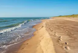 Pusta plaża nudystów z falami obmywającymi brzeg i śladami stóp na piasku.