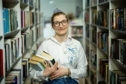 Bibliotekarka z okularami trzyma stos książek. W tle regały pełne literatury. Czy wakat co to znaczy?