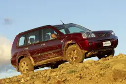 Czerwony Nissan X-Trail ubrudzony błotem podjeżdża pod skaliste wzniesienie.