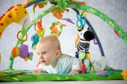 Niemowlę leży na macie edukacyjnej z zabawkami: żyrafą, zebrą i ptaszkiem.