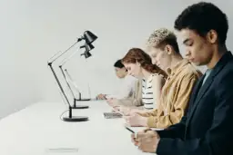 Młodzi ludzie pracują przy biurkach. Zastanawiają się, czy staż jest płatny, skupieni na swoich laptopach i telefonach.