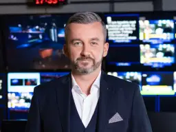 Krzysztof Skórzyński, elegancki mężczyzna w garniturze, stoi na tle ekranów. Ile ma lat? Tego nie wiemy, ale wygląda na doświadczonego dziennikarza.