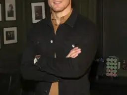 Sam Heughan w czarnej kurtce dżinsowej, z rękami skrzyżowanymi na piersi. W tle zdjęcia i ekspres do kawy.