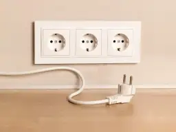 Biały kabel z wtyczką leży na drewnianym blacie, obok gniazdek elektrycznych w ścianie.