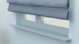 Szara roleta rzymska częściowo zasłania okno z białym parapetem.