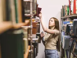 Młoda kobieta w bibliotece szuka inspiracji, zastanawiając się, jakie gatunki literackie są najbardziej poszukiwane przez wydawców w 2026.