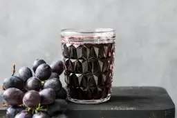 Szklanka ciemnego soku z winogron obok kiści świeżych winogron.
