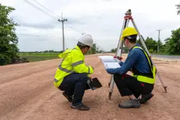 Dwóch geodetów w kamizelkach odblaskowych i kaskach analizuje plany na budowie. Zastanawiają się, ile bierze geodeta za wytyczenie działki.