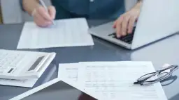 Osoba analizuje dokumenty, porównując umowę o pracę a umowę zlecenie, korzystając z laptopa i kalkulatora.