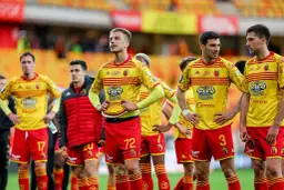 Zawodnicy Jagiellonii Białystok w żółto-czerwonych strojach stoją na boisku, gotowi do gry.