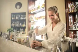 Uśmiechnięta barmanka przygotowuje drinka, ucząc się, jak zostać barmanem. Za nią półki z alkoholem.