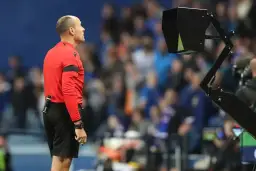 Sędzia w czerwonej koszulce czeka na decyzję z wozu VAR. Tłum kibiców na trybunach.