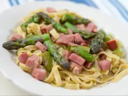Pyszne spaghetti ze szparagami i kawałkami szynki na białym talerzu.