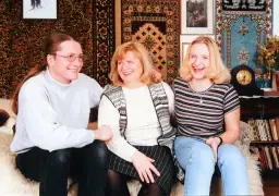Trzy uśmiechnięte osoby siedzą na kanapie: mężczyzna z długimi włosami i kobieta w środku, a obok nich młoda kobieta.
