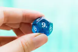 Palec i kciuk trzymają niebieską, dwudziestościenną kostkę do gry z widoczną cyfrą 9. To symbol numerologiczna 9, oznaczająca zakończenie cyklu.