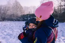 Mama w różowej czapce i granatowej kurtce tuli dziecko w nosidełku. Zima w pełni, śnieg wokół. Jak ubrać dziecko do nosidełka zimą? Warstwowo i ciepło!