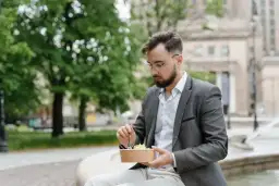 Mężczyzna w okularach i marynarce robi sobie przerwę w pracy, jedząc sałatkę na ławce w parku.