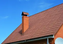 Ceglany komin z metalowym daszkiem, obrobiony blachą, na tle brązowego dachu i błękitnego nieba.