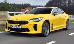 Żółty Kia Stinger GT pędzi po drodze, otoczony zielenią i domami.