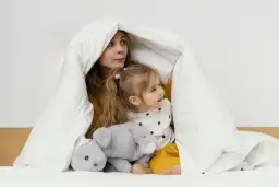 Mama i dziecko bawią się pod białą kołdrą, która przypomina przytulne kołderki dla niemowląt. Obok leży szary pluszowy króliczek.