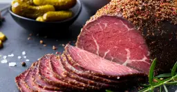Pokrojone plastry pastrami, przyprawione pieprzem, obok ogórków konserwowych. Zastanawiasz się pastrami co to? To aromatyczne, peklowane i wędzone mięso wołowe.