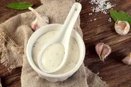 Crema de salsa de ajo en un cuenco blanco con una cuchara, rodeada de ajos y hojas verdes sobre una mesa de madera rústica.