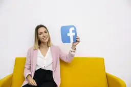 Uśmiechnięta kobieta na żółtej kanapie trzyma niebieski symbol Facebooka. To idealny przykład na marketing szeptany w social mediach.