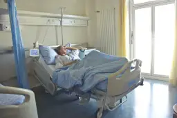 Pacjentka w szpitalnym łóżku, zastanawia się, zwolnienie lekarskie ile płatne. Jasne okno i zasłony.