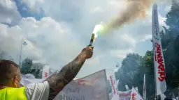 Dym z flary rozświetla niebo nad demonstracją związków zawodowych. W tle białe flagi z napisem "Solidarność".