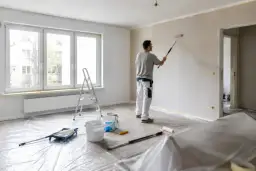 Malowanie pokoju: mężczyzna wałkiem odświeża ściany. Na podłodze folia ochronna, drabina i wiaderka z farbą.