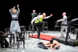 Scena teatralna z kobietą w szarej sukni, mężczyzną w kamizelce odblaskowej i leżącym na bieżni. Czy to symbol tego, ile trwają przerwy w teatrze?
