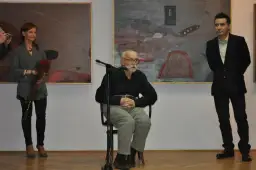 Konrad Wieniawa-Narkiewicz siedzi przy mikrofonie, obok niego stoi kobieta z kwiatami i mężczyzna w garniturze.