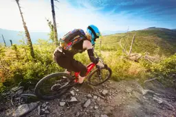 Kobieta w kasku i ochraniaczach zjeżdża rowerem po kamienistym single track w Polsce. Słoneczny dzień, górzysty krajobraz.