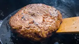 Jugosa hamburguesa de ternera dorándose en sartén, con vapor ascendiendo y una espátula de madera lista para darle la vuelta.