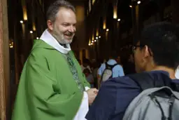 Uśmiechnięty ksiądz w zielonej szacie wita młodego mężczyznę. Warto wiedzieć, jak się zwracać do biskupa, ale i do każdego kapłana z szacunkiem.