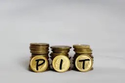 Trzy stosy monet, na których widnieją litery P, I, T. Symbolizują one rozliczenie podatku PIT.