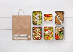 Co do grama catering: pięć pudełek z jedzeniem, torba i butelka wody na białym drewnianym stole.