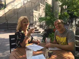 Tom Logan, mąż Majki Jeżowskiej, uśmiecha się do zdjęcia, siedząc przy stoliku na zewnątrz z drinkiem.