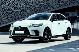 Biały Lexus RX 500h z czarnymi felgami stoi przed nowoczesnym budynkiem o geometrycznej fasadzie.
