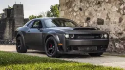 Mroczny Dodge Challenger Demon, z potężną maską i agresywnym grillem, stoi przed kamienną ścianą.