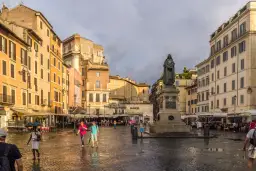Pomnik na placu Campo de' Fiori w Rzymie, otoczony kolorowymi budynkami i ludźmi spacerującymi po mokrym bruku.