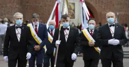 Rycerze Kolumba w garniturach i maskach maszerują z flagą.
