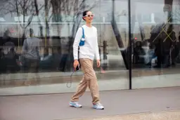 Mujer sonriente camina con un suéter blanco y un pantalón beige de carga, luciendo gafas de sol blancas y un bolso azul.
