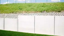 Solidny mur oporowy z betonowych płyt, zabezpieczający zielone zbocze. Nad nim ażurowa warstwa, a dalej trawnik i niebo.