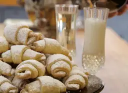 Stos puszystych rogalików krucho drożdżowych, posypanych cukrem pudrem, obok kieliszków z napojem.